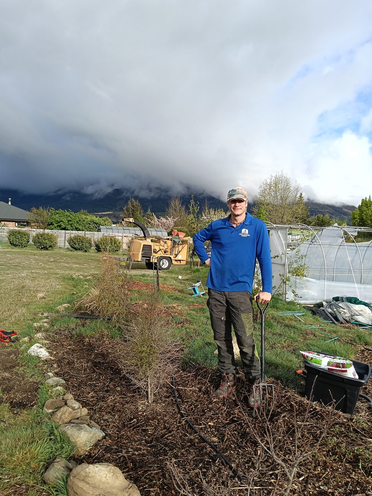 An arborist with a garden fork beside a freshly planted and mulched bed, a wood chipper and tunnel house behind